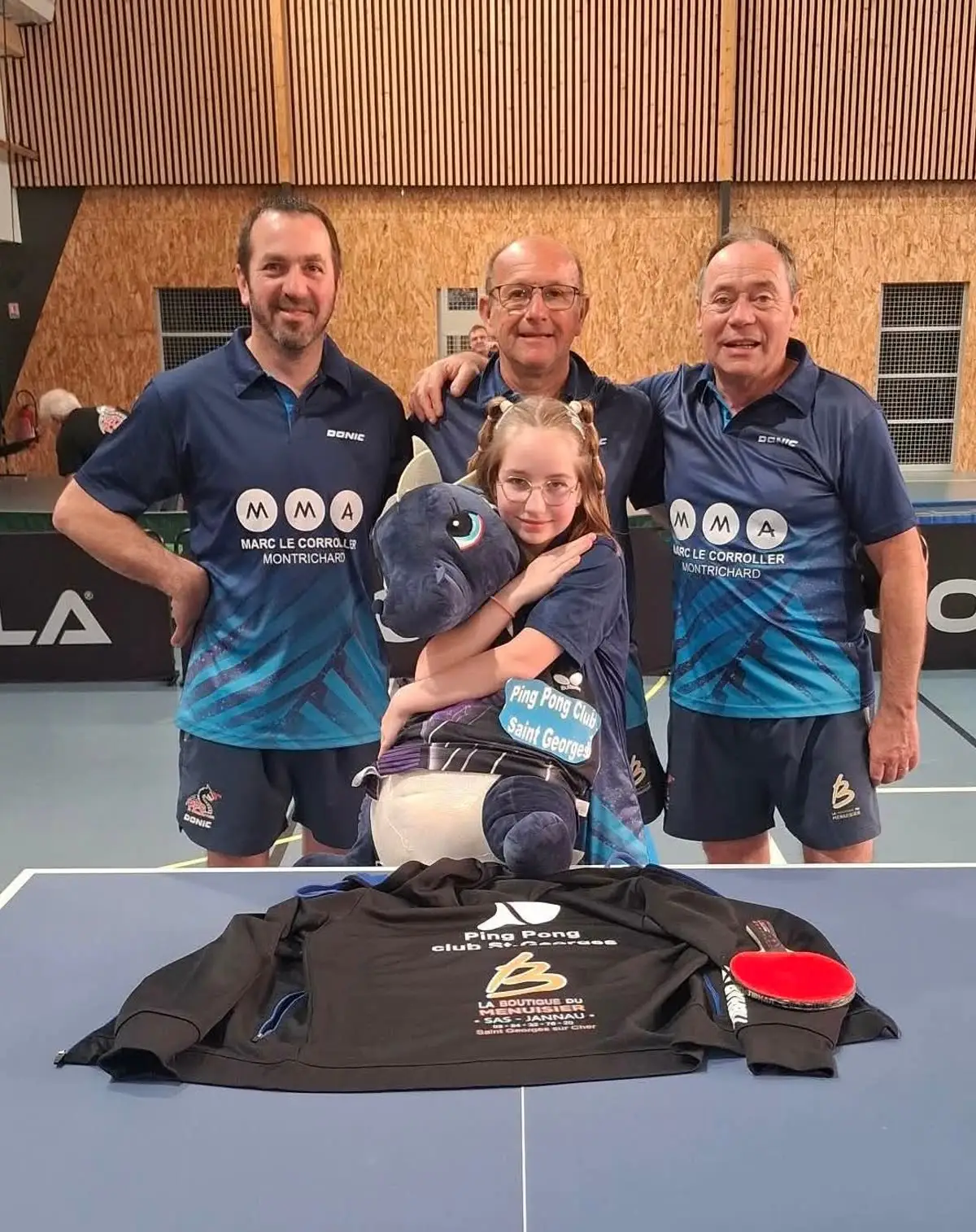 Trois joueurs adultes en maillot du club entourant une jeune licenciée qui tient la mascotte dragon, derrière un maillot floqué posé sur une table.